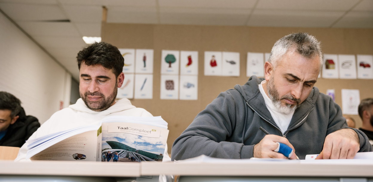 Twee nieuwkomers zitten gebogen over hun lesboeken tijdens de Nederlandse les